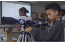 A young girl adjusts a large blue telescope in a classroom.